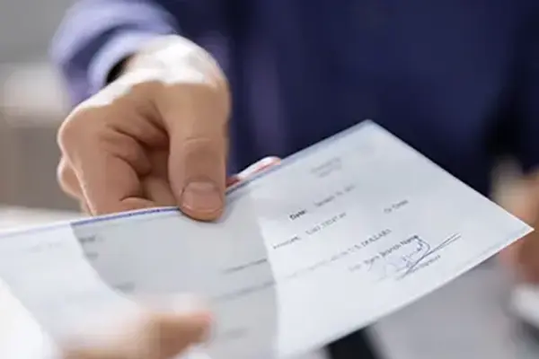 This image shows a person in a blue shirt handing a check or a document to another person in an office or clinical setting. The focus is on the outstretched hand and the check/document.