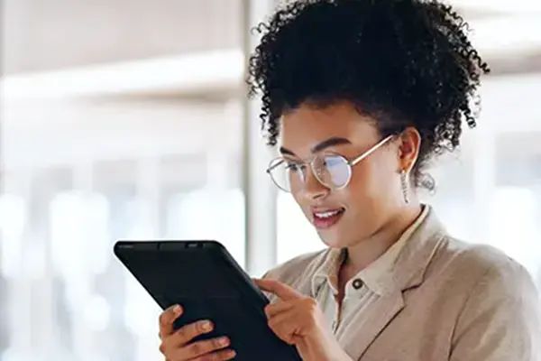 This image shows a woman with curly hair and glasses, looking at a digital tablet with interest. She appears to be in a modern, bright indoor setting, possibly a cafe or a coworking space.