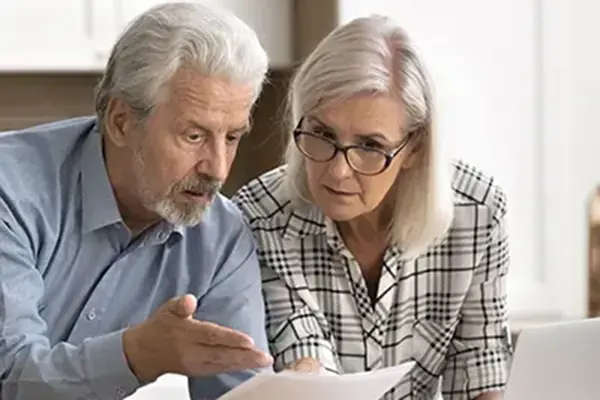 This image shows an elderly couple discussing documents at a kitchen table, with a laptop open in front of them. They appear to be engaged in a serious conversation, possibly reviewing financial or legal papers.